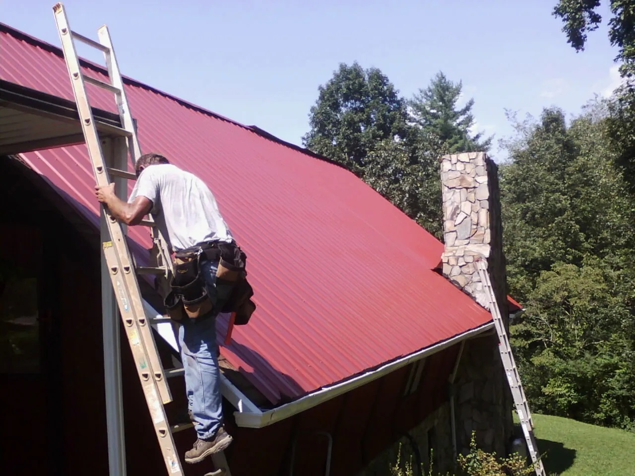 Roofer on a ladder beside a red metal roof and stone chimney — Soffit & Fascia Repair by Roofing Brothers in Nevada City